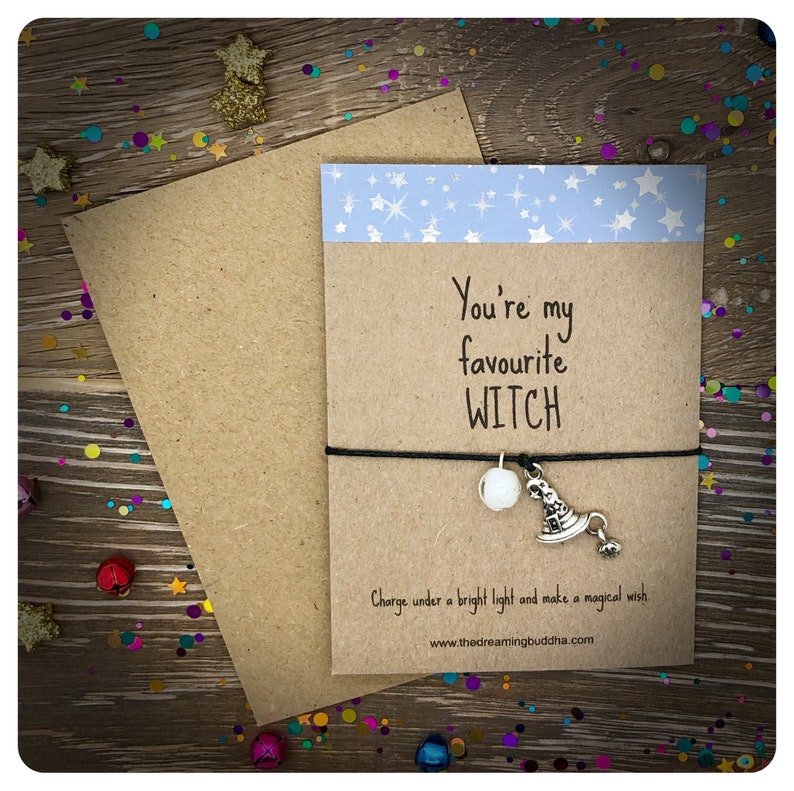 May include: A brown card with the text "You're my favourite WITCH" and a black string bracelet with a white bead and a silver charm of a witch's hat. The card has a blue and white background with stars.