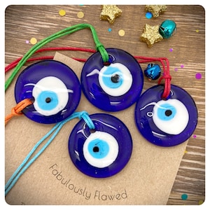 May include: Four blue glass evil eye charms with white and blue eyes and black pupils, each charm is attached to a colourful string. The charms are on a brown paper background with the text "Fabulously Flawed".