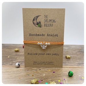 May include: Orange string anklet with a silver bee charm. The anklet is on a card that says "The Dreaming Buddha Handmade Anklet Follow your own path. Made in Lancashire England www.thedreamingbuddha.com"