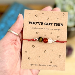 May include: A red string bracelet with three gemstone beads: tiger's eye, carnelian, and clear quartz. The bracelet is displayed on a brown card with the text "YOU'VE GOT THIS" and star illustrations. The card also lists the gemstone types.