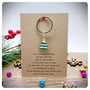 May include: A brown card with a gold key ring attached to a green and white Christmas tree charm. The card has a handwritten message that reads: "I'm a little pine tree as you can see. Life in the forest is still very new to me. I'm thankful for your hard work in helping me to grow. So I'm sending you this little card just to let you know."