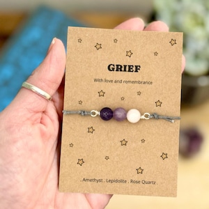 May include: A bracelet featuring amethyst, lepidolite, and rose quartz beads on a grey cord. The bracelet is presented on a card with the word "GRIEF" and the text "With love and remembrance". The card is decorated with star illustrations.