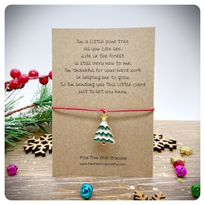 May include: A red string bracelet with a gold charm of a green and white Christmas tree. The card attached to the bracelet reads "I'm a little pine tree as you can see. Life in the forest is still very new to me. I'm thankful for your hard work in helping me to grow. So I'm sending you this little card just to let you know."  The card also says "Pine Tree Wish Bracelet" and "www.thedreamingbuddha.com".