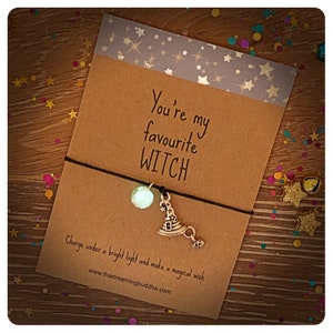 May include: A brown card with the text "You're my favourite WITCH" and a black string bracelet with a silver witch hat charm and a clear bead. The card also has the text "Charge under a bright light and make a magical wish. www.thedreamingbuddha.com"