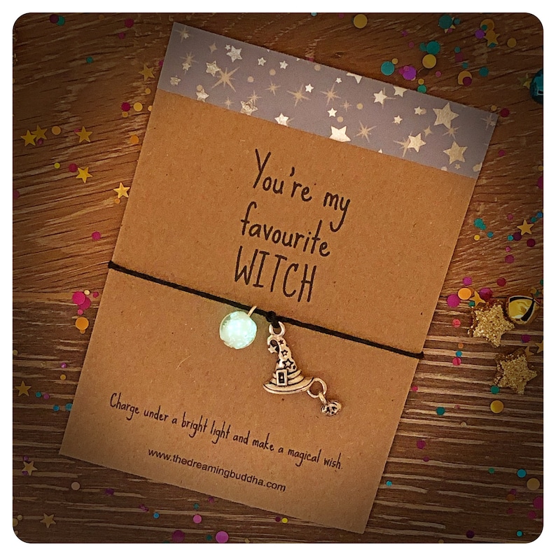 May include: A brown card with the text "You're my favourite WITCH" and a black string bracelet with a silver witch hat charm and a clear bead. The card also has the text "Charge under a bright light and make a magical wish. www.thedreamingbuddha.com"