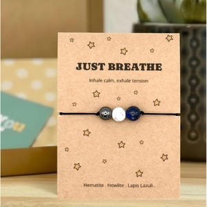May include: A bracelet featuring three beads—a metallic hematite, a white-veined howlite, and a blue lapis lazuli—on a black cord. The display card reads "JUST BREATHE, Inhale calm, exhale tension" and lists the stones: Hematite, Howlite, Lapis Lazuli.