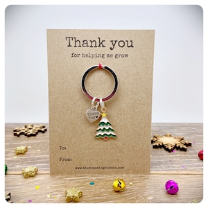 May include: A festive Christmas keychain featuring a green and white tree charm and a heart charm with the words "Thank You". The keychain is on a brown card with the text "Thank you for helping me grow".