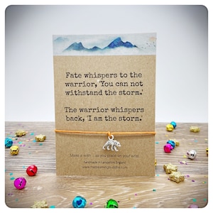 May include: A brown card with a quote about a warrior and a storm. The card is tied with a string and has a silver bear charm. The card is on a wooden surface with colorful confetti.