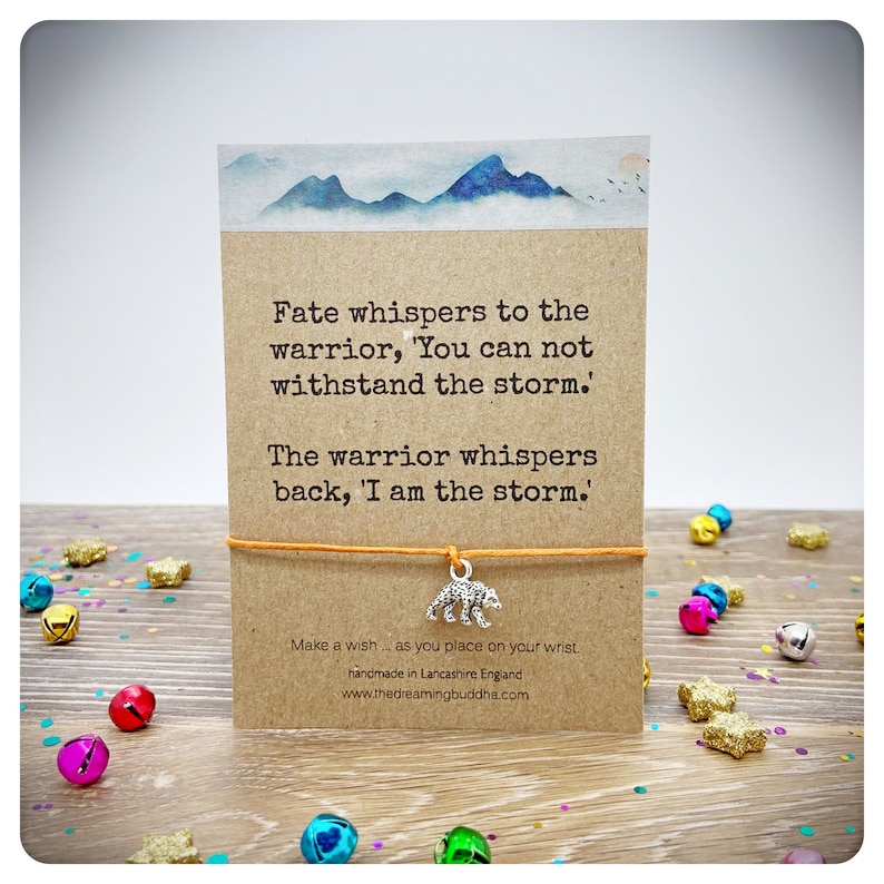 May include: A brown card with a quote about a warrior and a storm. The card is tied with a string and has a silver bear charm. The card is on a wooden surface with colorful confetti.