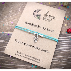 May include: A turquoise blue string anklet with a silver dreamcatcher charm and silver beads. The anklet is displayed on a card with the text "The Dreaming Buddha Handmade Anklet Follow your own path. Made in Lancashire England www.thedreamingbuddha.com"