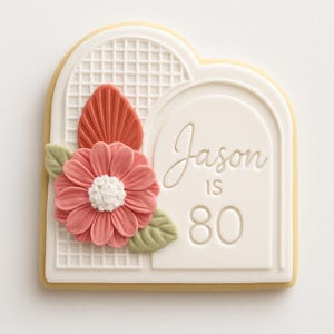 May include: A decorated cookie in the shape of a white arch with a lattice design. It features a coral-colored flower, a leaf, and the text "Jason is 80" in script.