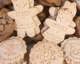 May include: Assortment of decorated sugar cookies, including two gingerbread girl cookies with the names "Clara" and "Audrey" on them, and other cookies with holiday themes like a snowflake and a Christmas tree.