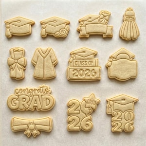 May include: A collection of graduation-themed cookies in various shapes, including mortarboards, diplomas, and graduation gowns. Some cookies feature the text "Class of 2026" and "congrats GRAD". The cookies are light brown and arranged on a light-colored surface.