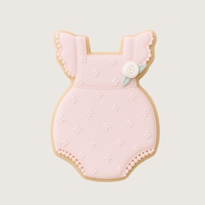 May include: A light pink onesie-shaped cookie with a floral design. The cookie has a scalloped edge and is decorated with small flower-shaped details. A white flower with green leaves is placed on the front.