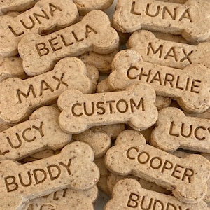 May include: A collection of bone-shaped dog biscuits with names and the word "CUSTOM" imprinted on them. The biscuits are light brown and appear to be homemade. Names include Luna, Bella, Max, Charlie, Lucy, Buddy, and Cooper.