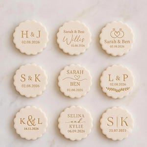 May include: Nine round, scalloped-edge cream-colored cookies featuring gold-colored printed text and graphics. Designs include initials like H & J, S & K, L & P, K & L, S | K, and names like Sarah & Ben, Willis, and Selina and Kylie with dates.