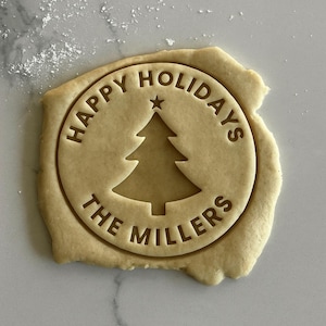 May include: A circular cookie dough stamp featuring a central Christmas tree icon topped with a star. The text "HAPPY HOLIDAYS" is arched across the top, and "THE MILLERS" is arched across the bottom, all pressed into the dough.