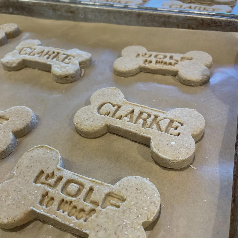 Puede incluir: Primer plano de galletas caseras para perros con forma de hueso. Las galletas son de color marr&oacute;n claro y tienen nombres como "Clarke" y "Wolf to Woof" grabados. Est&aacute;n dispuestas sobre una bandeja para hornear forrada con papel de hornear.