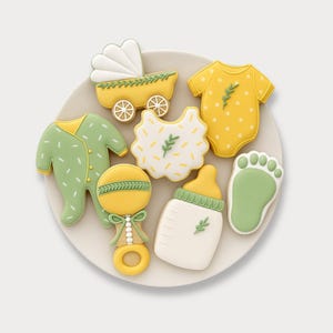 Puede incluir: Una colección de galletas con temática de bebé dispuestas en un plato blanco. Las galletas incluyen un cochecito amarillo, un pelele verde, un pelele amarillo con lunares blancos, un babero, un sonajero, un biberón y una huella, todos decorados con detalles verdes.
