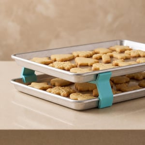 May include: Two rectangular silver metal baking sheets stacked vertically and held apart by four light blue plastic clips. Each tray is filled with various light brown, baked holiday-shaped cookies, including stars, gingerbread people, and candy canes.