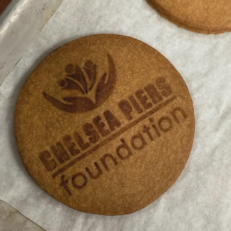 Puede incluir: Una galleta redonda de color marr&oacute;n claro con las palabras "CHELSEA PIERS foundation" y un logotipo grabado en la superficie. La galleta est&aacute; sobre una superficie blanca.