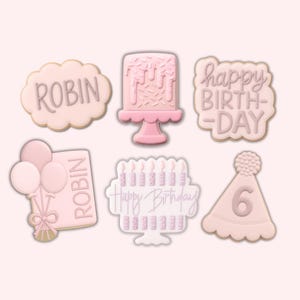 Pode incluir: Uma cole&ccedil;&atilde;o de biscoitos cor-de-rosa com tema de anivers&aacute;rio. Os biscoitos incluem formas como um bolo, bal&otilde;es, um chap&eacute;u de festa com o n&uacute;mero 6 e biscoitos com as palavras "ROBIN" e "Happy Birthday".