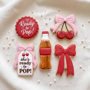 May include: Five decorated sugar cookies: a red bottle cap with white text "Ready to Pop!", a pair of cherries with a light pink bow, a white tag with a pink bow and "she's ready to POP!" text, a brown soda bottle, and a large red bow.