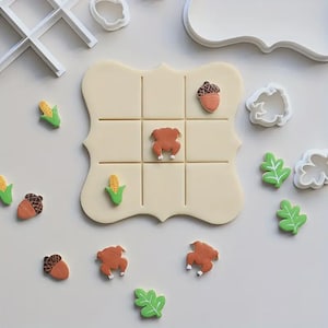 May include: A Thanksgiving-themed tic-tac-toe game with a beige board and grid. The game pieces include corn, acorns, turkeys, and green leaves. White cookie cutters and a tic-tac-toe grid are also present, suggesting a baking theme.