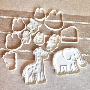 Puede incluir: Un conjunto de cortadores de galletas de pl&aacute;stico blanco en varias formas, incluyendo una jirafa, un elefante, un conejo y una nube. Los cortadores est&aacute;n dispuestos sobre una superficie beige y blanca a rayas. Ideales para galletas con tem&aacute;tica animal.
