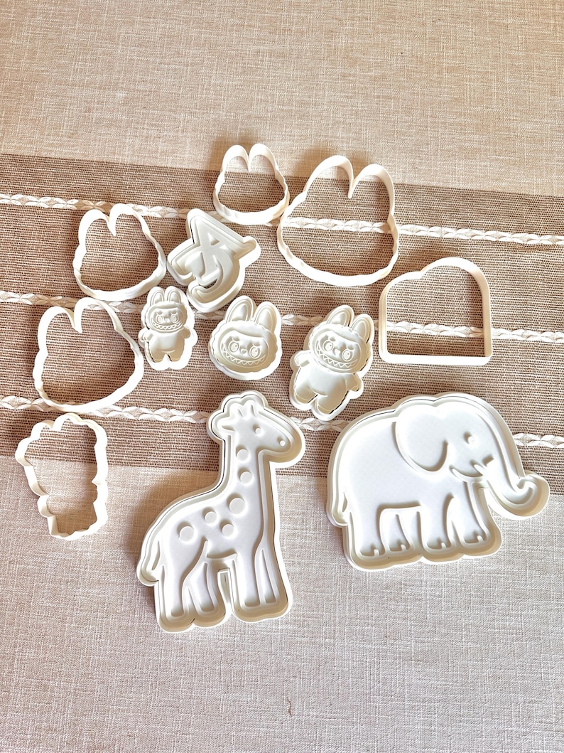 Puede incluir: Un conjunto de cortadores de galletas de pl&aacute;stico blanco en varias formas, incluyendo una jirafa, un elefante, un conejo y una nube. Los cortadores est&aacute;n dispuestos sobre una superficie beige y blanca a rayas. Ideales para galletas con tem&aacute;tica animal.