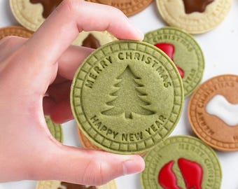 May include: A hand holding a green cookie with a Christmas tree design and the words "MERRY CHRISTMAS" and "HAPPY NEW YEAR" embossed around the edge. Other cookies with various designs and colors are in the background.