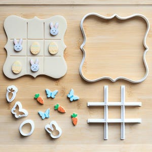 May include: A collection of Easter-themed cookie cutters and a decorated tic-tac-toe game. The game features bunny and egg designs. Additional cutters include butterflies, carrots, and a bunny head. The items are white and pastel-colored.