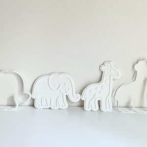Puede incluir: Cuatro cortadores de galletas blancos con forma de animales: un hipop&oacute;tamo, un elefante, una jirafa y una segunda jirafa. El elefante tiene detalles, mientras que los otros son contornos. Los objetos est&aacute;n sobre una superficie blanca.