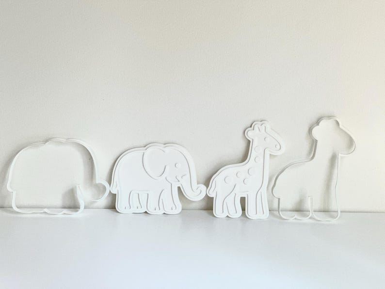 Puede incluir: Cuatro cortadores de galletas blancos con forma de animales: un hipop&oacute;tamo, un elefante, una jirafa y una segunda jirafa. El elefante tiene detalles, mientras que los otros son contornos. Los objetos est&aacute;n sobre una superficie blanca.