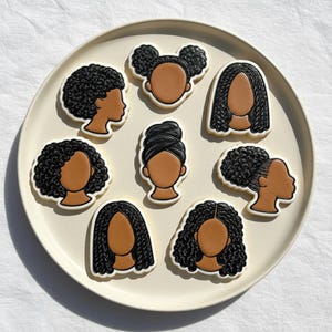 May include: A collection of eight cookies arranged on a cream-colored plate. Each cookie features a brown face with different black hairstyles, including braids, buns, and afros. The cookies are outlined in white.