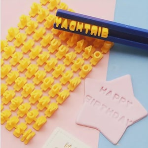 May include: A set of yellow alphabet stamps with a blue handle, used to imprint letters. The word "YAOHTAIB" is stamped. A pink star-shaped item is stamped with "HAPPY BIRTHDAY".