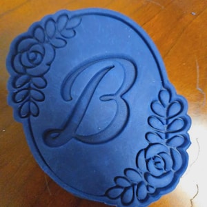 Puede incluir: Un cortador de galletas azul marino en forma ovalada, con la letra "B" en el centro. Los bordes est&aacute;n decorados con dise&ntilde;os florales y de hojas, creando un borde decorativo. El cortador est&aacute; hecho de un material flexible.
