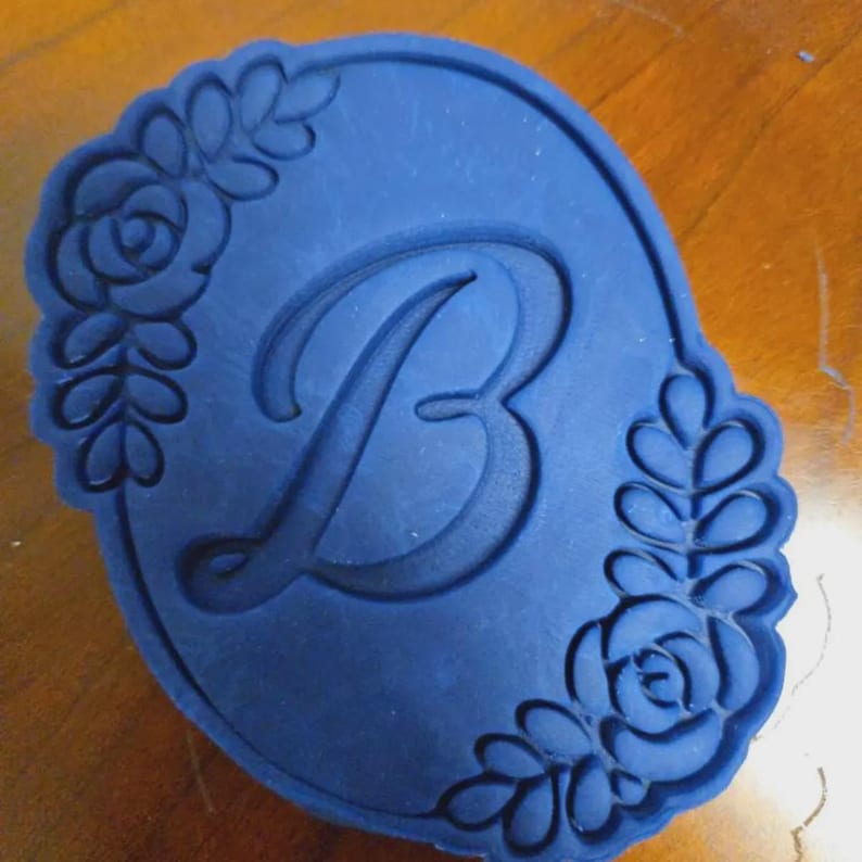 Puede incluir: Un cortador de galletas azul marino en forma ovalada, con la letra "B" en el centro. Los bordes est&aacute;n decorados con dise&ntilde;os florales y de hojas, creando un borde decorativo. El cortador est&aacute; hecho de un material flexible.