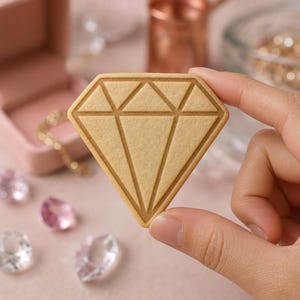 May include: A light tan, diamond-shaped cookie featuring embossed geometric lines that mimic the facets of a cut gemstone. The cookie has a smooth, matte surface and is held up to display its distinct, symmetrical, angular shape.