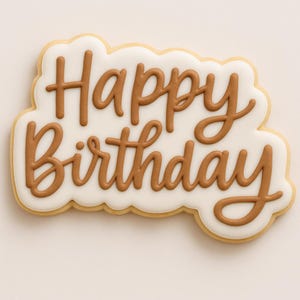 May include: A decorated sugar cookie with the words "Happy Birthday" in brown icing. The cookie has a white base with a scalloped edge and a light brown border. The lettering is in a cursive style.