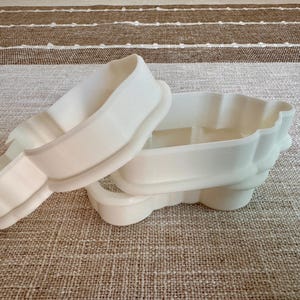 May include: Two white plastic cookie cutters. One is a rectangular shape with rounded edges, and the other is a similar shape with a scalloped edge. The cutters are on a neutral-colored fabric surface.