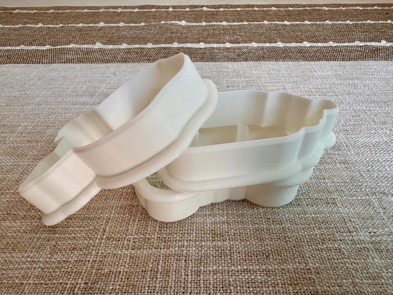 May include: Two white plastic cookie cutters. One is a rectangular shape with rounded edges, and the other is a similar shape with a scalloped edge. The cutters are on a neutral-colored fabric surface.