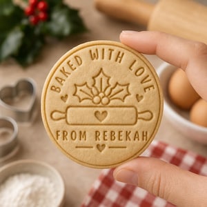 May include: A round, light tan cookie featuring an embossed design of holly leaves, berries, and a rolling pin with a heart in the center. The text reads BAKED WITH LOVE FROM REBEKAH, with small heart accents above and below the rolling pin.