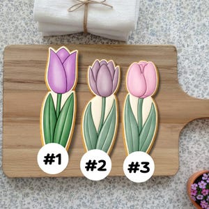 May include: Three tulip-shaped cookies with different colored petals: purple, lavender, and pink. Each cookie has green leaves and a white stem, set on a wooden cutting board. The cookies are labeled with numbers in white circles.