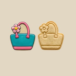 May include: Two cookie cutters shaped like handbags. One is decorated with teal, pink, and yellow icing, featuring a flower accent. The other is a plain, unadorned cookie cutter. The handbag design includes a handle and a flower.