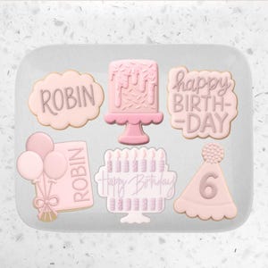 Pode incluir: Uma cole&ccedil;&atilde;o de biscoitos cor-de-rosa com tema de anivers&aacute;rio. Os biscoitos incluem um bolo, um chap&eacute;u de festa com o n&uacute;mero 6, um biscoito com o nome "Robin", um biscoito com bal&otilde;es e um biscoito que diz "Happy Birthday".