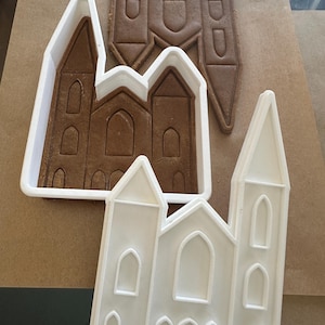 Puede incluir: Una galleta de jengibre con forma de iglesia y dos cortadores de galletas de pl&aacute;stico blanco con forma de iglesia. La galleta es marr&oacute;n y tiene detalles arquitect&oacute;nicos. Los cortadores son blancos y tienen el mismo dise&ntilde;o de iglesia.
