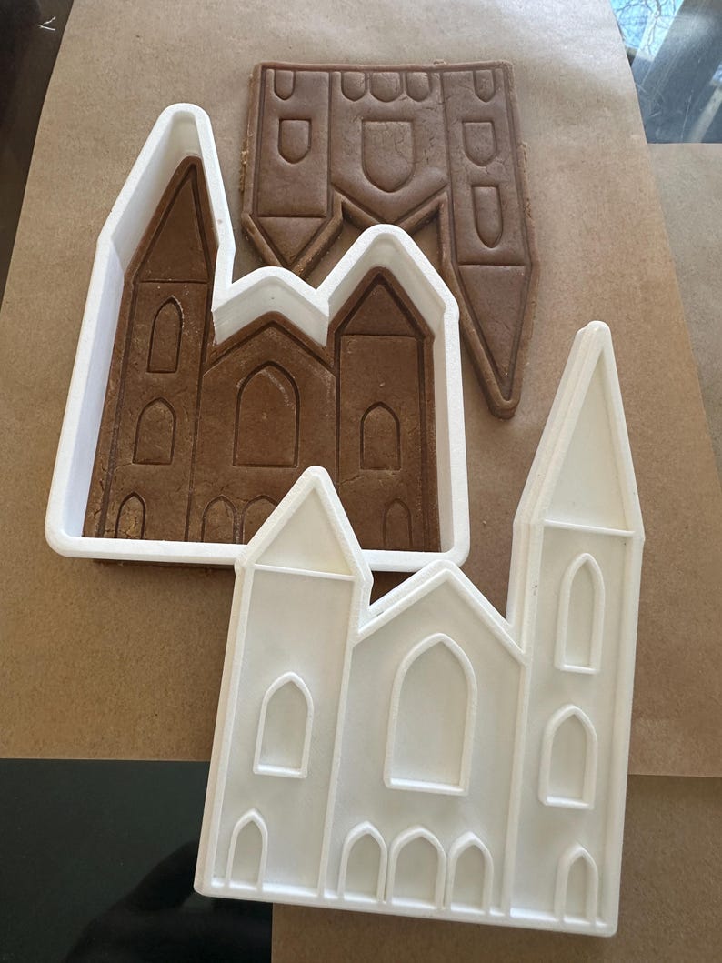 Puede incluir: Una galleta de jengibre con forma de iglesia y dos cortadores de galletas de pl&aacute;stico blanco con forma de iglesia. La galleta es marr&oacute;n y tiene detalles arquitect&oacute;nicos. Los cortadores son blancos y tienen el mismo dise&ntilde;o de iglesia.