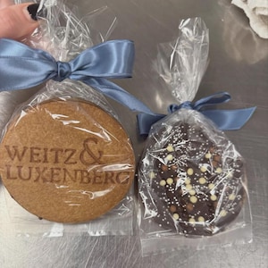 Puede incluir: Dos galletas envueltas, una redonda y otra ovalada, est&aacute;n atadas con una cinta de raso azul claro. La galleta redonda es de color marr&oacute;n claro con el texto "WEITZ & LUXENBERG" en relieve. La galleta ovalada es de color marr&oacute;n oscuro y est&aacute; cubierta de chispas blancas y marrones claras.
