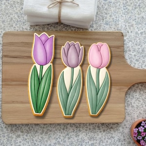 May include: Three tulip-shaped cookies with purple, lavender, and pink petals, green leaves, and a light-colored stem. The cookies are on a wooden cutting board. A stack of white towels and a small pot of flowers are in the background.